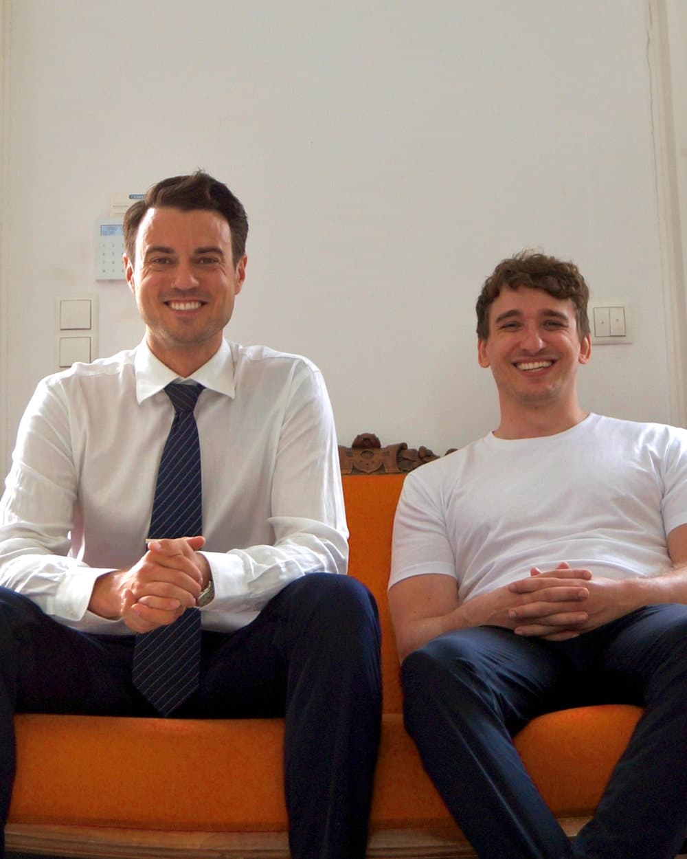 Johannes Freyer und Torben Lange sitzen lachend nebeneinander auf einem orangefarbenen Sofa
